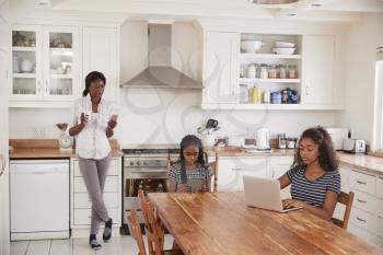 Mother Uses Phone As Daughters Sit At Table Doing Homework