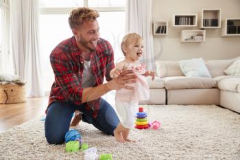 Young father helping toddler daughter walk in sitting room