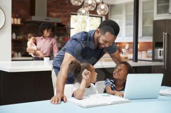 Father Helps Children With Homework Whilst Mother With Baby Uses Laptop In Kitchen