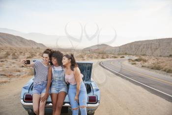 Three Female Friends Posing For Selfie Sitting In Trunk Of Classic Car On Road Trip