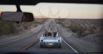 Group Of Friends On Road Trip Driving Classic Convertible Car Viewed Through Windshield Of Following Vehicle