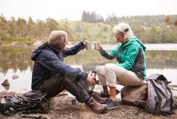 Senior couple sitting by a lake drinking coffee during camping holiday making a toast with their mugs, Lake District, UK