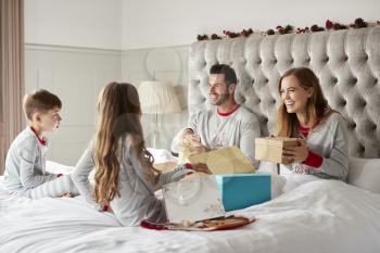 Excited Children Sitting On Parents Bed At Home As Family Open Gifts On Christmas Day