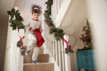 Excited Boy Wearing Pajamas Running Down Stairs Holding Stocking On Christmas Morning