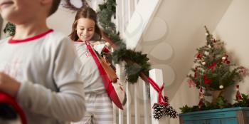 Two Excited Children Wearing Pajamas Running Down Stairs Holding Stockings On Christmas Morning