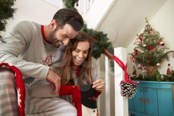 Excited Couple Wearing Pajamas Sitting On Stairs Looking Into Stockings On Christmas Morning