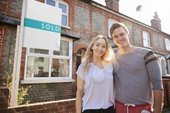 Portrait Of Excited Couple Standing Outside New Home With Sold Sign