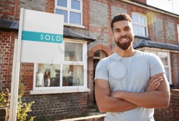 Portrait Of Excited Man Standing Outside New Home With Sold Sign