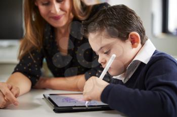 Close up of young female teacher sitting at desk with a Down syndrome schoolboy using a tablet computer in a primary school classroom, close up, side view