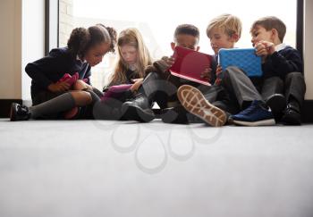 Low angle view of primary school friends sitting together in front of a window in a school corridor looking at tablet computers together