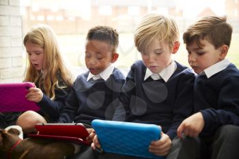 Four primary school children sitting on the floor in front of a window using tablet computers during break time, close up