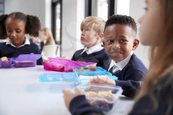 Smiling primary school kids sitting at a table eating their packed lunches together, selective focus