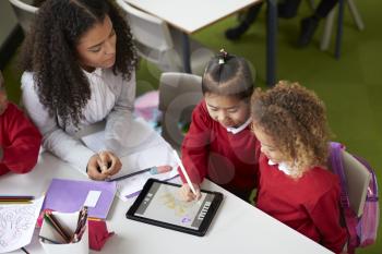 Elevated view of two girls using a tablet computer sitting at table in an infant school class a with female teacher helping them