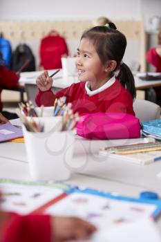 Close up of young Chinese schoolgirl wearing school uniform sitting at a desk in an infant school classroom, selective focus, vertical