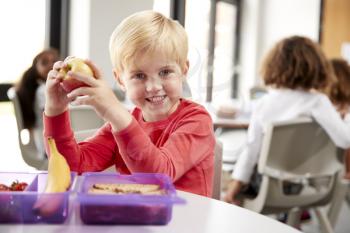 Young white schoolboy sitting at a table smiling and holding an apple in a kindergarten classroom during his lunch break, close up