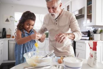 Young girl breaking an egg into cake mixture with her grandfather at the kitchen table, close up