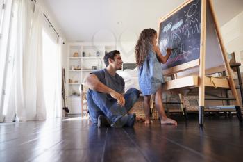 Hispanic dad sitting on the floor in sitting room watching his young daughter drawing on blackboard