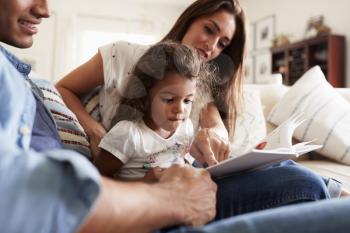 Hispanic couple and their young daughter sitting on the sofa reading a book together at home