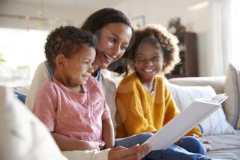 Close up of young mother sitting on a sofa in the living room reading a book to her two children, close up, side view