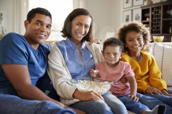 Young mixed race family sitting together on the sofa in their living room, eating popcorn and looking to camera, front view