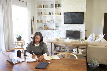 Happy middle aged mixed race woman sitting at table in her dining room making notes, elevated view