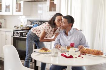 Young adult woman kissing her partner, sitting at the table in their kitchen for a romantic dinner, selective focus