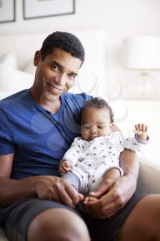 Close up of young adult black father sitting in an armchair holding his three month old baby son, smiling to camera, vertical