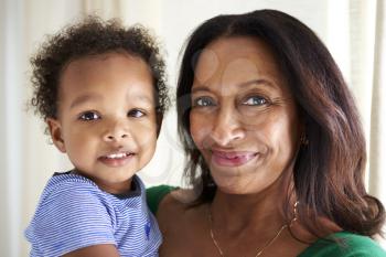 Happy mixed race middle aged grandmother holding her two year old grandson, both looking to camera smiling, close up, head and shoulders