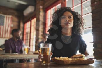 Woman Watching Game On Screen In Sports Bar Eating Burger And Fries