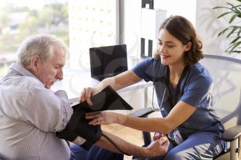 Nurse Wearing Scrubs In Office Checking Senior Male Patients Blood Pressure