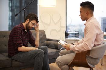 Unhappy Man Sitting On Couch Meeting With Male Counsellor In Office