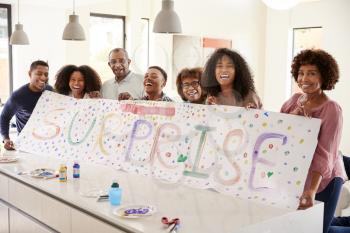 Three generation black family holding up a sign they’ve made for a surprise party at home