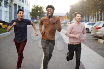 Three hip young adult male friends running for fun in a city road, front view, three quarter length