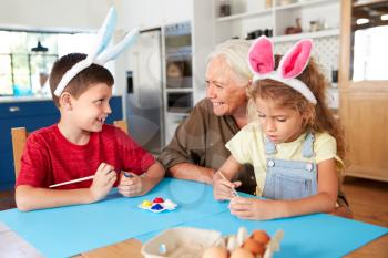 Grandmother With Grandchildren Wearing Rabbit Ears Decorating Easter Eggs At Home Together