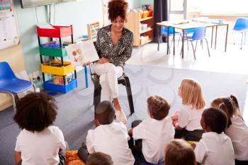 Female Teacher Reading Story To Group Of Elementary Pupils Wearing Uniform In School Classroom