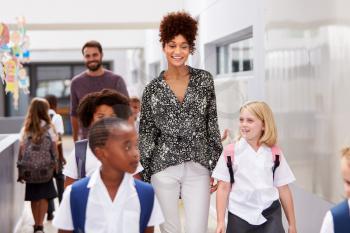 Teacher And Pupils Walking Along Corridor In Busy Elementary School Corridor