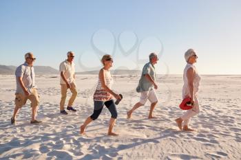 Side View Of Senior Friends Walking Along Sandy Beach On Summer Group Vacation