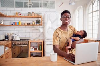 Portrait Of Multi-Tasking Father Holding Sleeping Baby Son And Working On Laptop Computer In Kitchen