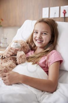 Portrait Of Girl Lying In Bed In Hospital Ward Hugging Teddy Bear