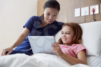 Female Nurse With Girl Lying In Hospital Bed Looking At Digital Tablet Together
