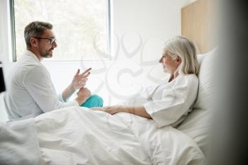 Doctor Wearing White Coat Visiting Mature Female Patient In Hospital Bed