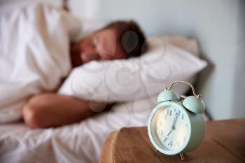 Mid adult man asleep in bed, alarm clock on the bedside table in the foreground, focus on foreground