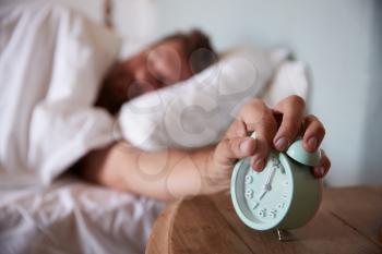 Mid adult man asleep in bed, reaching out to alarm clock on the bedside table in the foreground