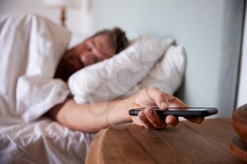 Mid adult man asleep in bed, reaching out to smartphone on the bedside table in the foreground