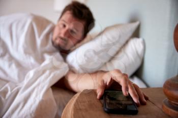 Mid adult man lying in bed, reaching out to smartphone on the bedside table in the foreground