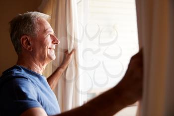 Smiling senior white man opening the curtains on a sunny morning, side view, close up