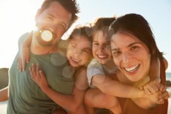 Mid adult white parents piggybacking their kids on a beach, smiling to camera, close up, lens flare