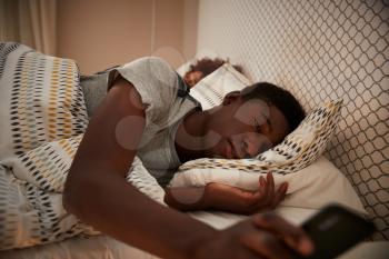 Millennial African American man half asleep in bed holding smartphone, his partner in the background, close up
