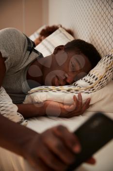 Millennial African American man half asleep in bed holding smartphone, his partner in the background, vertical