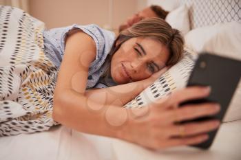 White woman half asleep in bed looking at smartphone, her partner sleeping in background, close up